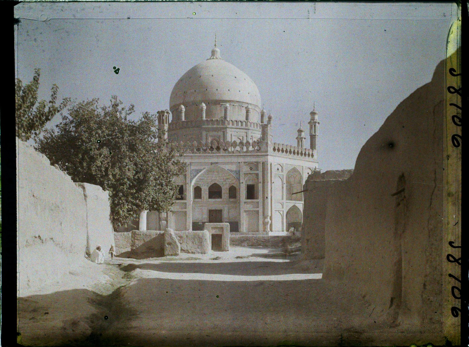 An early colour photograph of a domed white mausoleum with small minarets at its corners, seen down a dusty lane between mud walls, with a tree to the left and a seated figure in the shade.