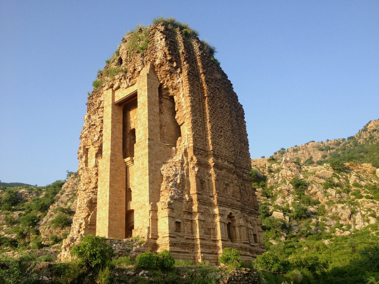 A tall ruined stone temple of several storeys with a tapering top, standing on a scrub-covered hillside under a clear sky.