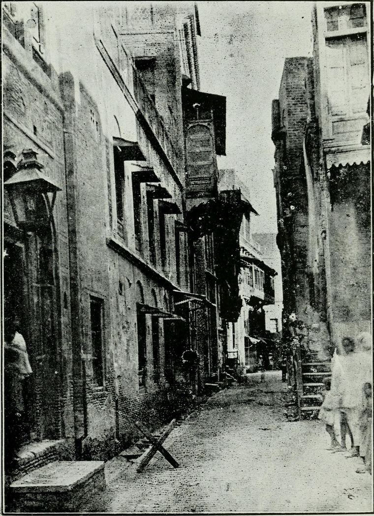 A narrow paved lane between tall brick houses with wooden balconies and awnings, a few children standing at the right