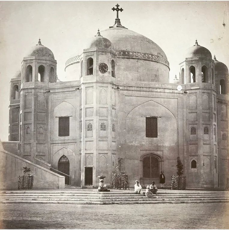 An octagonal Mughal tomb with a large central dome and domed corner towers, a cross fixed on top of the dome and a clock set into one tower, photographed in the nineteenth century with a few people sitting on the steps.