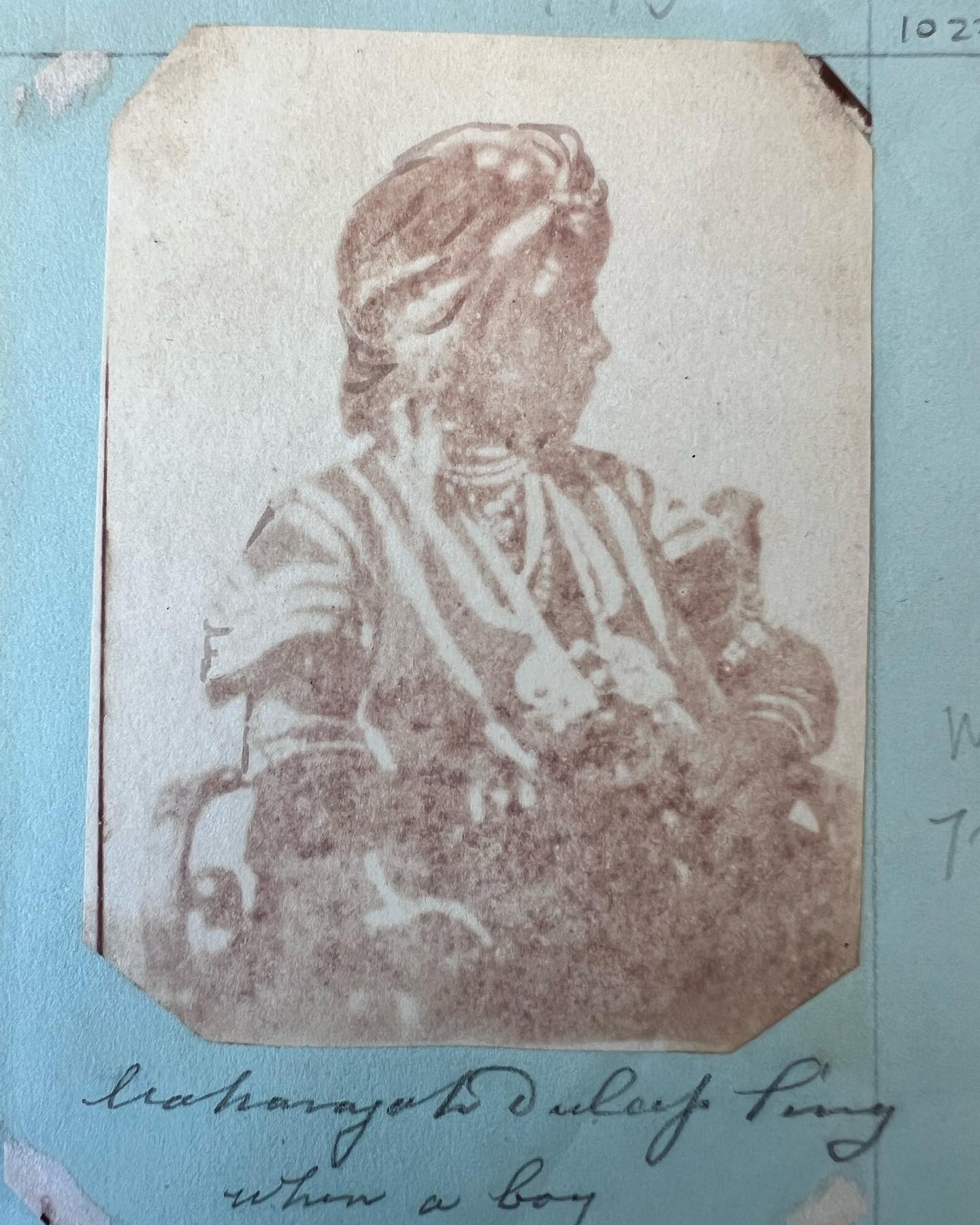 A faded reddish-brown photograph, mounted on a pale blue album page, of a boy in a turban and jewelled robes seated in profile, with a handwritten caption beneath it