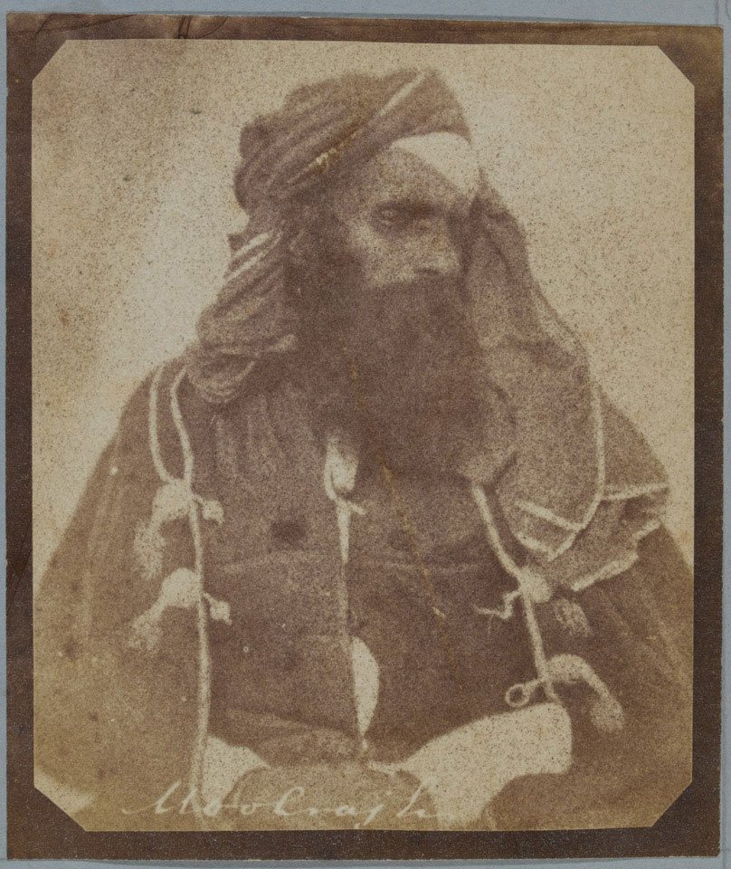 A faded sepia photograph of a bearded man in a turban and a heavy cloak with corded fastenings, looking to one side, with a name written along the bottom of the print