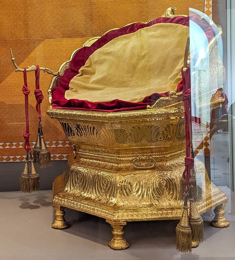 A low golden seat with a rounded back and lotus-petal base, upholstered in crimson velvet, in a glass museum case
