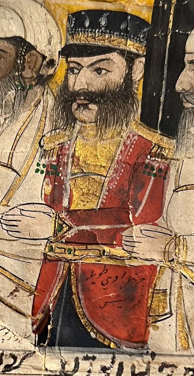 Detail of a painting of a court: a black-bearded man in a red European-style coat with gold facings and a black cap, hands folded at his waist, with Persian writing across the skirt of his coat.