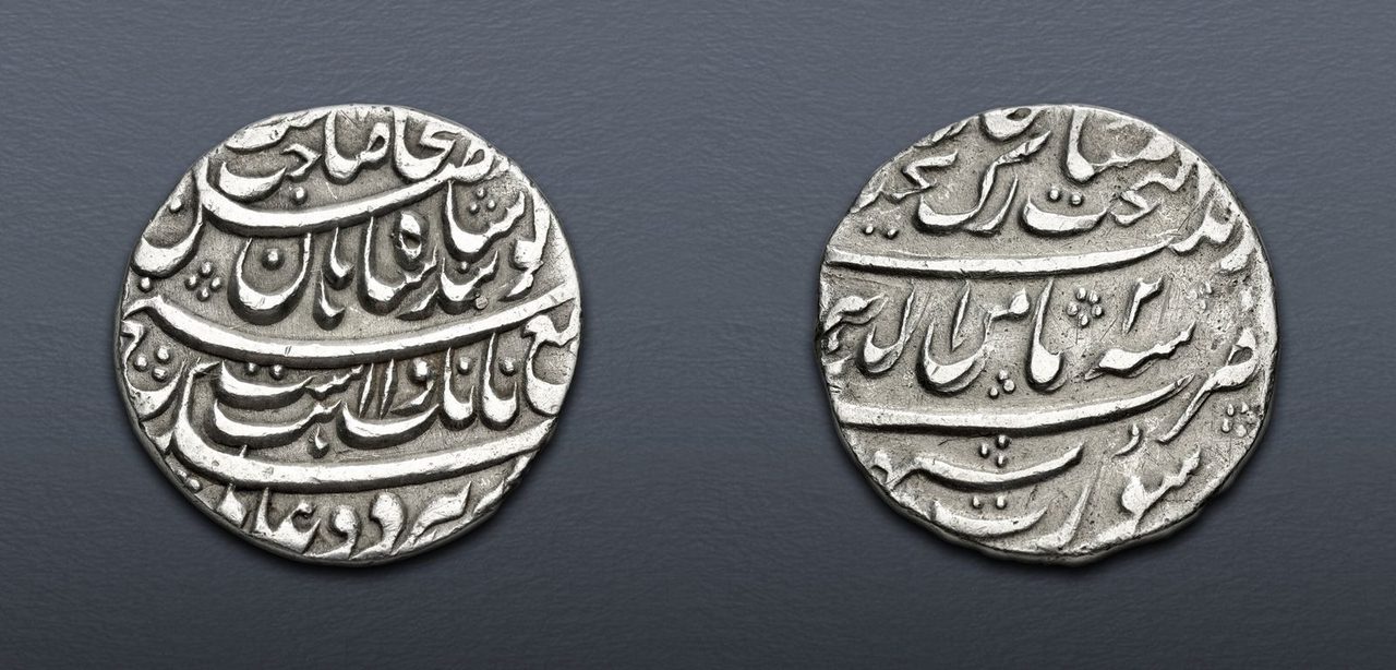 Both faces of a silver rupee side by side on a grey ground, each covered in raised Persian script set in bands.