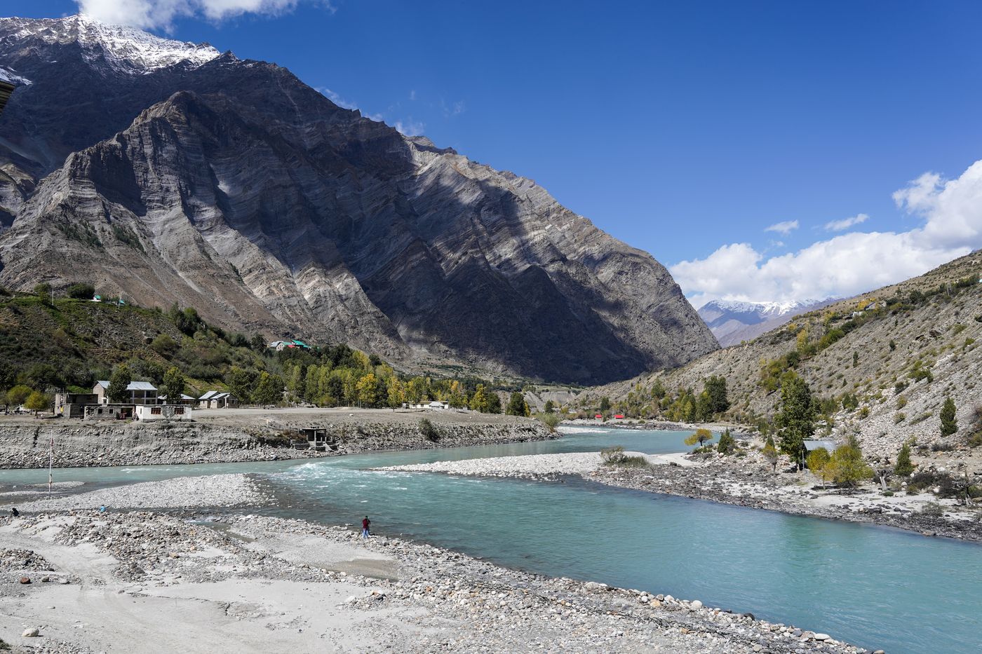 Two clear turquoise streams meeting on a wide gravel bed beneath a huge grey mountain face, with snow peaks behind and a deep blue sky.
