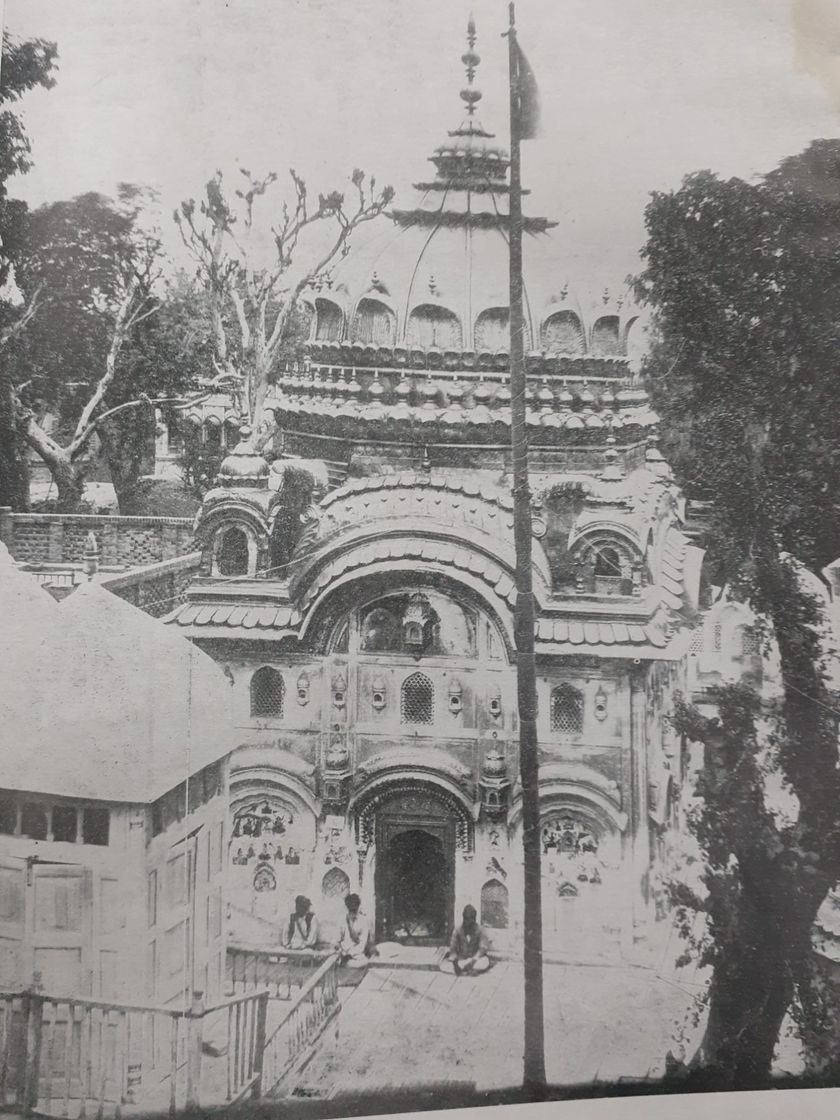 An old photograph of an ornate domed shrine with carved arches and painted panels on its front, a tall flagpole beside it, and three men seated at the entrance