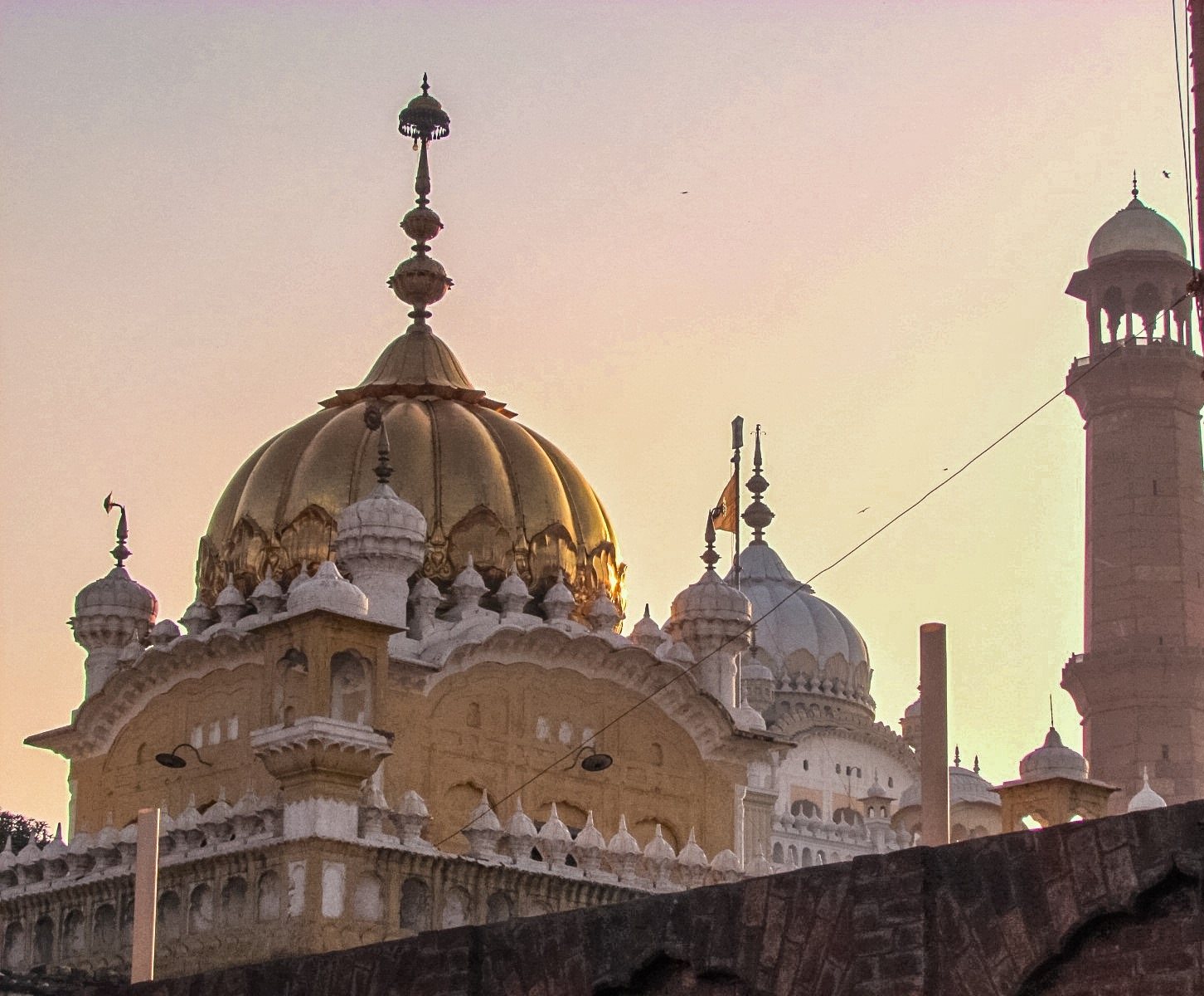 At dusk, the gilded ribbed dome of a gurdwara in the foreground, the white domes of a larger memorial behind it, and a tall red sandstone minaret with a domed top on the right.