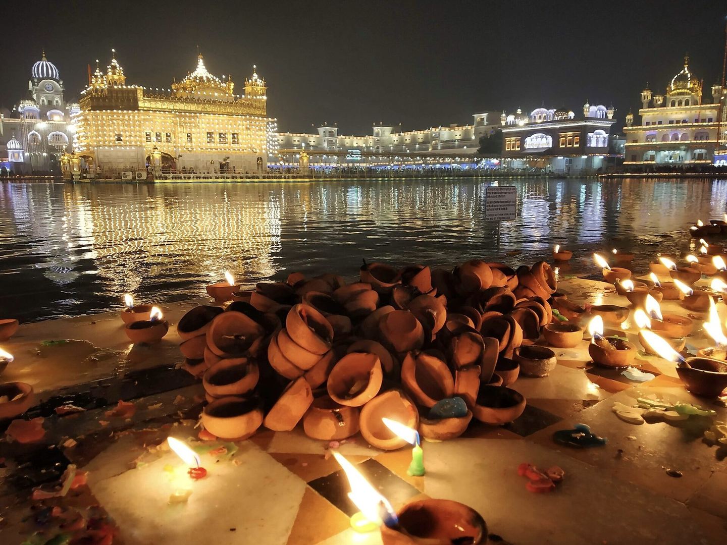 Night at the Golden Temple in Amritsar: the gilded shrine and the buildings round the pool strung with lights, their reflections on the dark water, and in front a heap of clay lamps on the marble walkway, some of them burning.