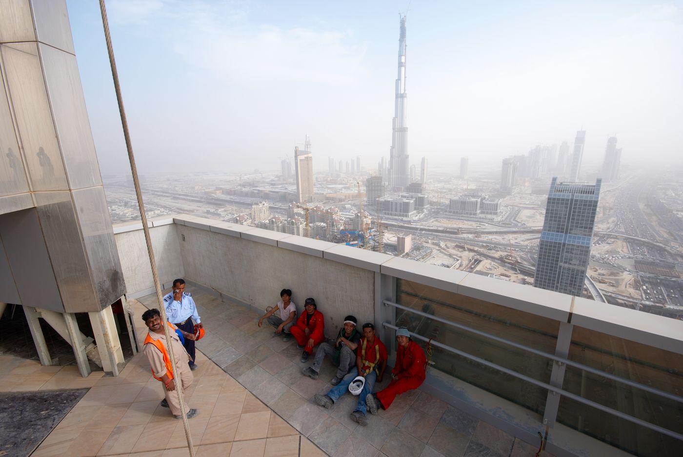 Five workers sit against the parapet of a high rooftop terrace, three of them in red, while two men stand at the left, one in an orange safety vest and one in a pale blue uniform shirt; behind them the unfinished Burj Dubai tower rises over a hazy city.