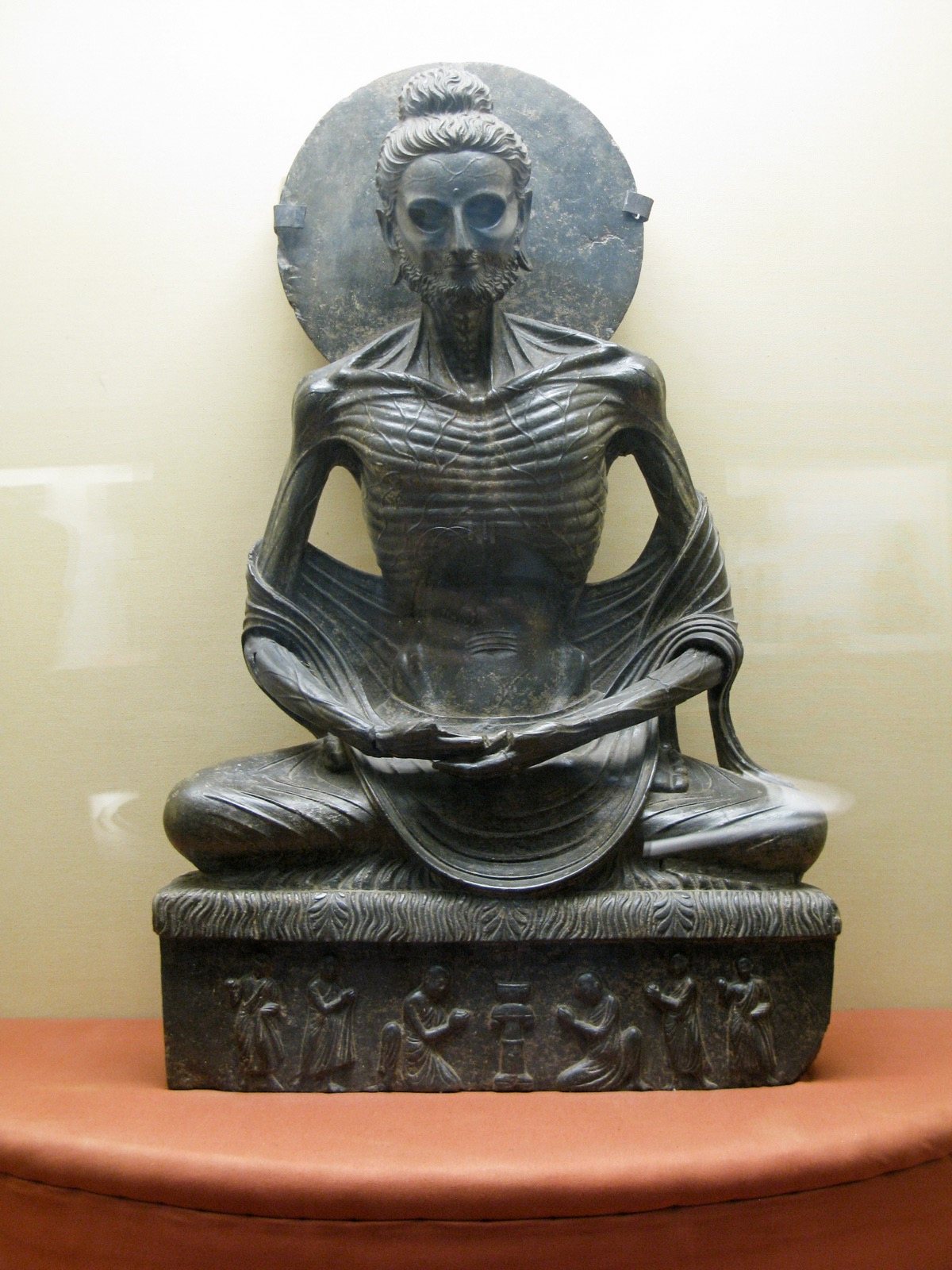 A dark grey stone sculpture of a seated, cross-legged figure so emaciated that every rib and vein shows, with a round halo behind the head and a carved base below
