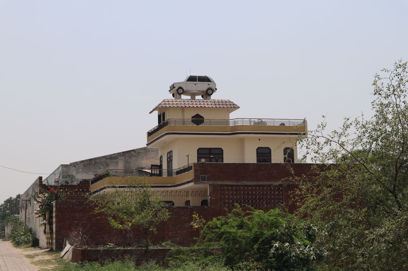 A cream-coloured house of two storeys behind a brick wall, with a white model car standing on a plinth on its roof against a hazy sky.