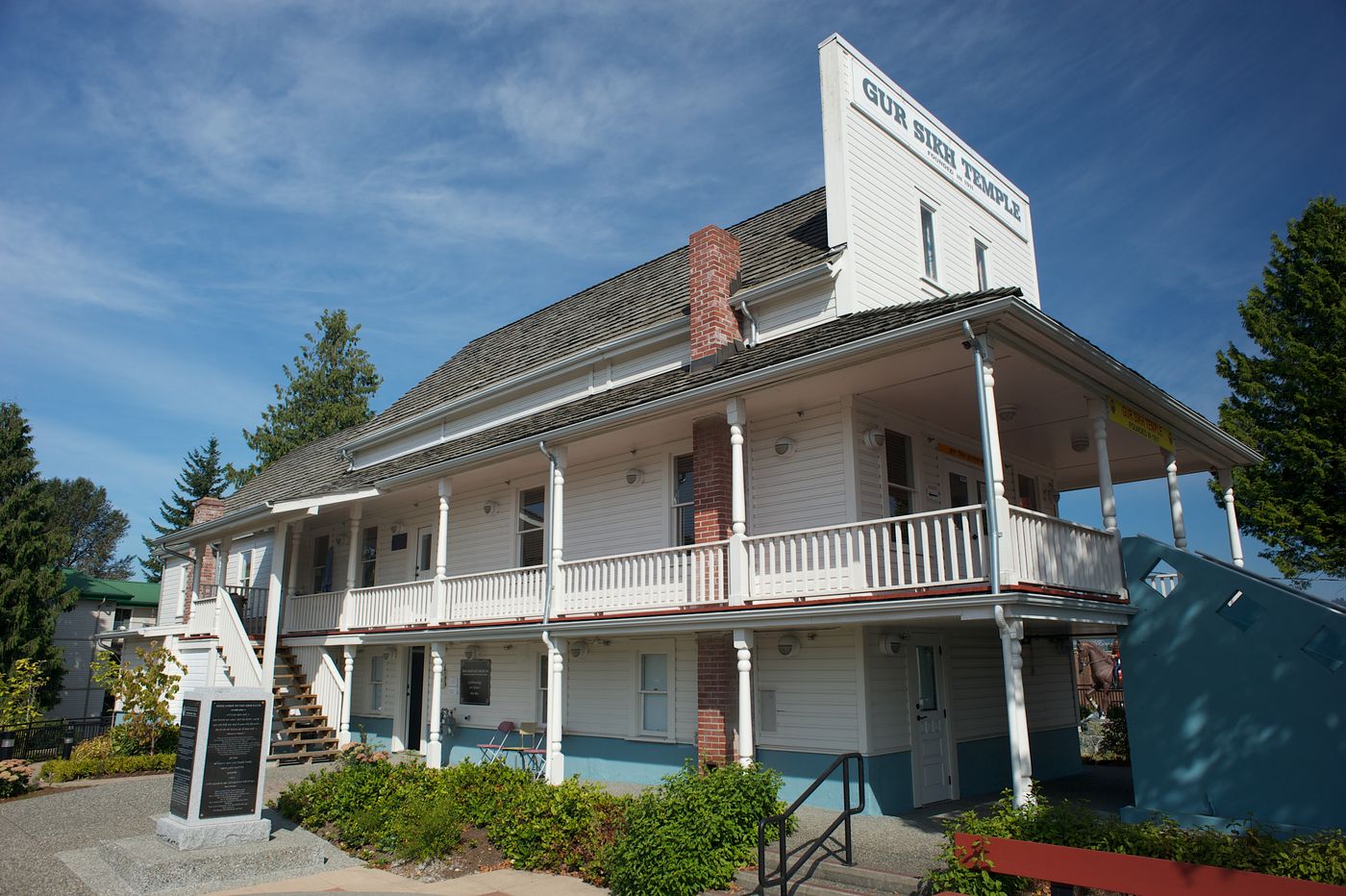 A white two-storey wooden building with a red-and-white balcony on two sides and a tall false front lettered Gur Sikh Temple, on a sunny day, with flower beds in front.
