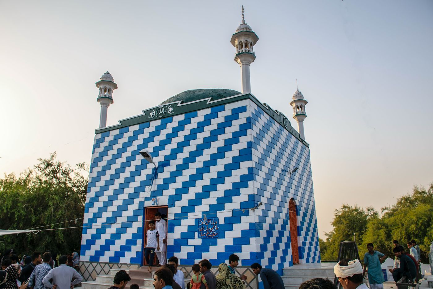 A square tomb faced in blue and white tiles laid in a stepped zigzag, with a low green dome and slender white minarets at its corners, and visitors gathered on the steps around it.