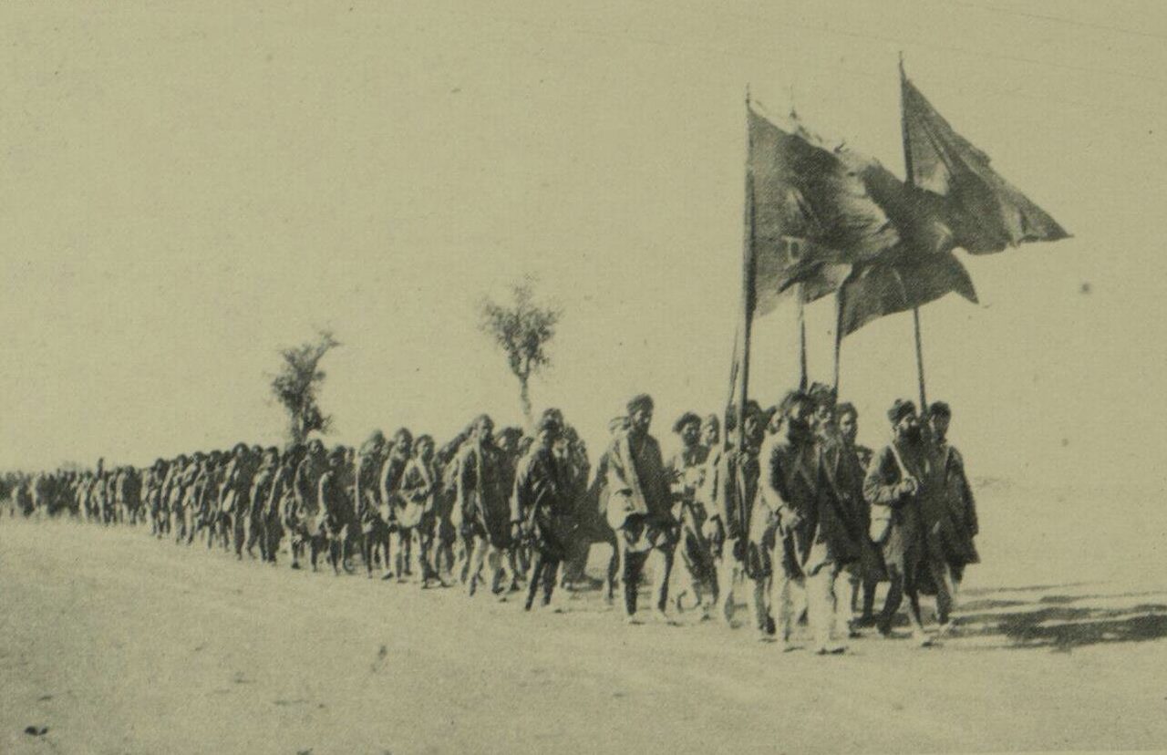 A long column of turbaned men walking along a flat, dusty road, three large flags on tall poles carried at its head and a couple of thin trees behind.