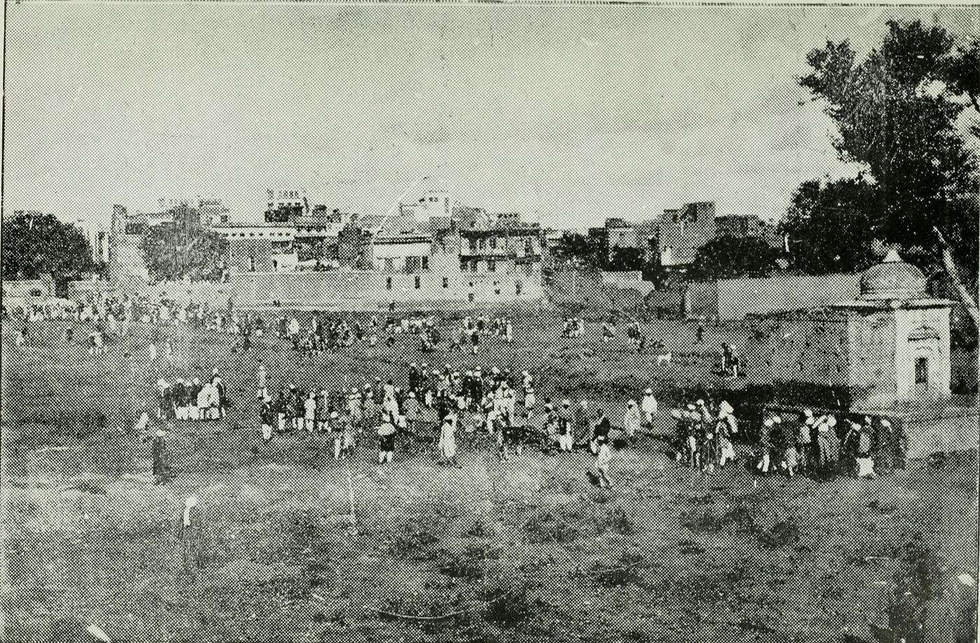 The Jallianwala Bagh in 1919 or 1920: rough open ground with groups of people, walls and houses behind, a small domed shrine at the right.