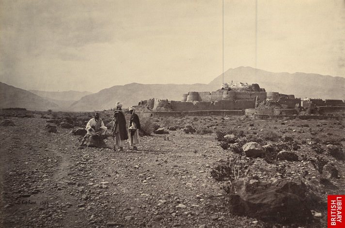 Old photograph of a large walled fort with round towers and a tall central building, on a low rise before a line of bare hills, with three men in turbans standing and sitting on the stony plain in front.