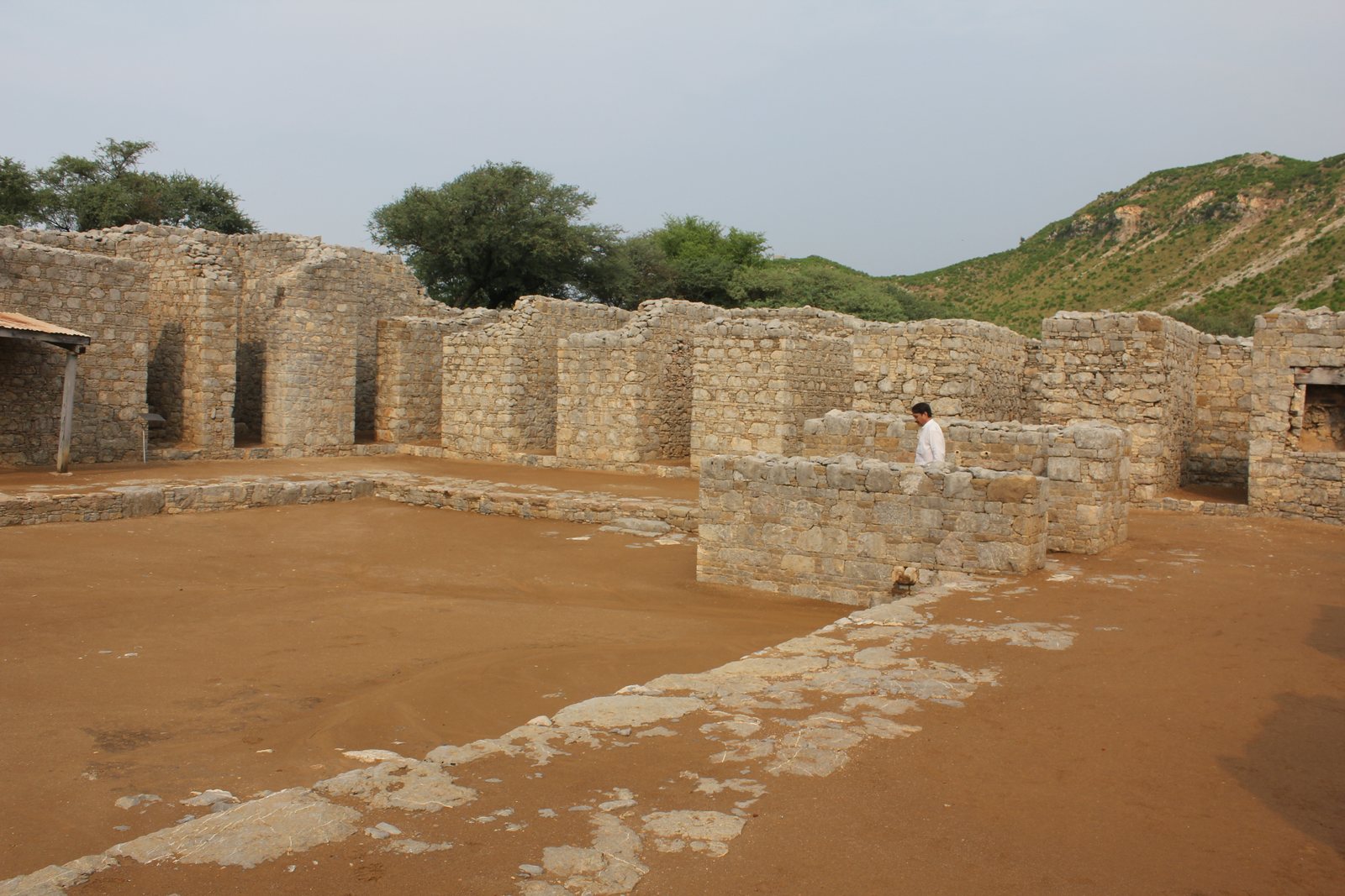 A courtyard of packed earth surrounded on three sides by the stone walls of small rooms with doorways, a hillside behind