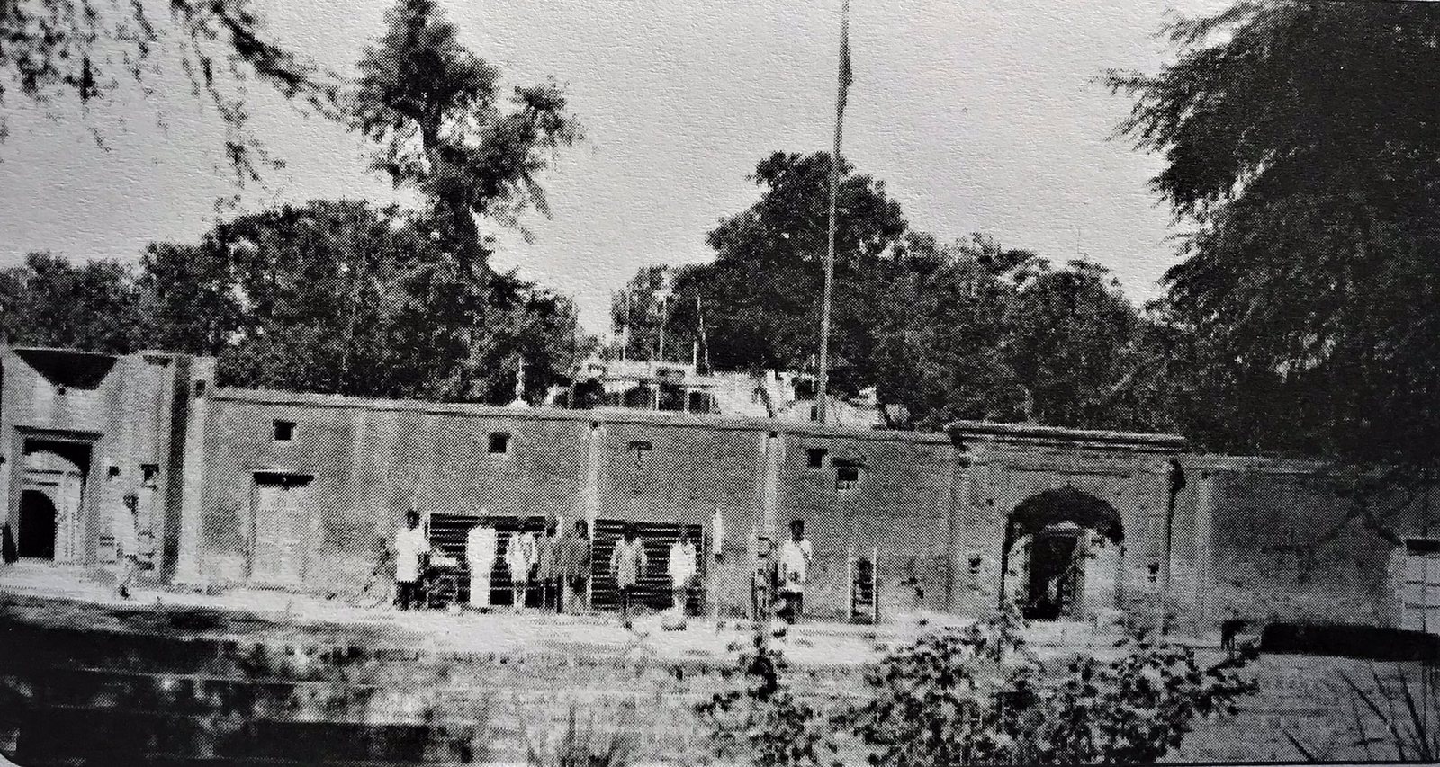 An old black-and-white photograph of a long, low compound wall with an arched gateway, a tall flagpole and a group of men standing before it, with trees and a building rising behind