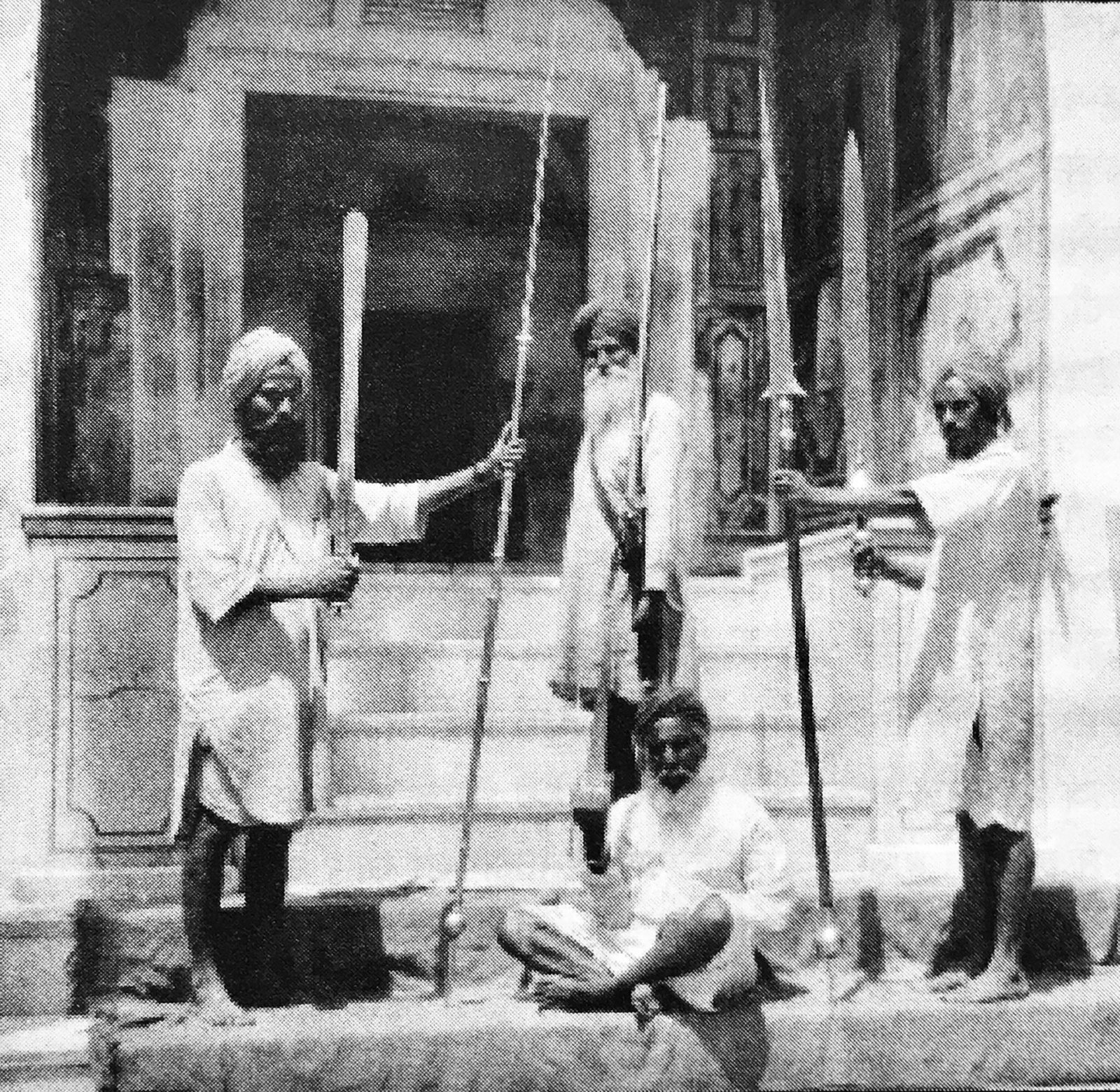 A black-and-white photograph of four Sikh men on the steps of a shrine holding up long swords, spears and a very long matchlock, one of them seated in front