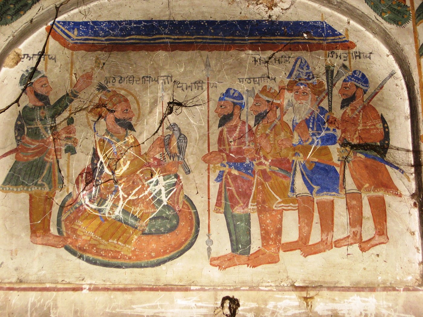 A cracked and faded wall painting under a painted blue canopy: a seated figure holding a hawk, a standing attendant at left, and five standing men with spears and swords at right, each with a line of Gurmukhi above his head