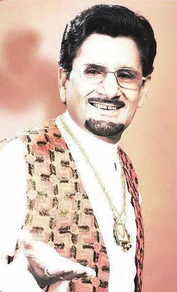 Portrait photograph of Kuldeep Manak in a patterned jacket, one hand raised as he sings.