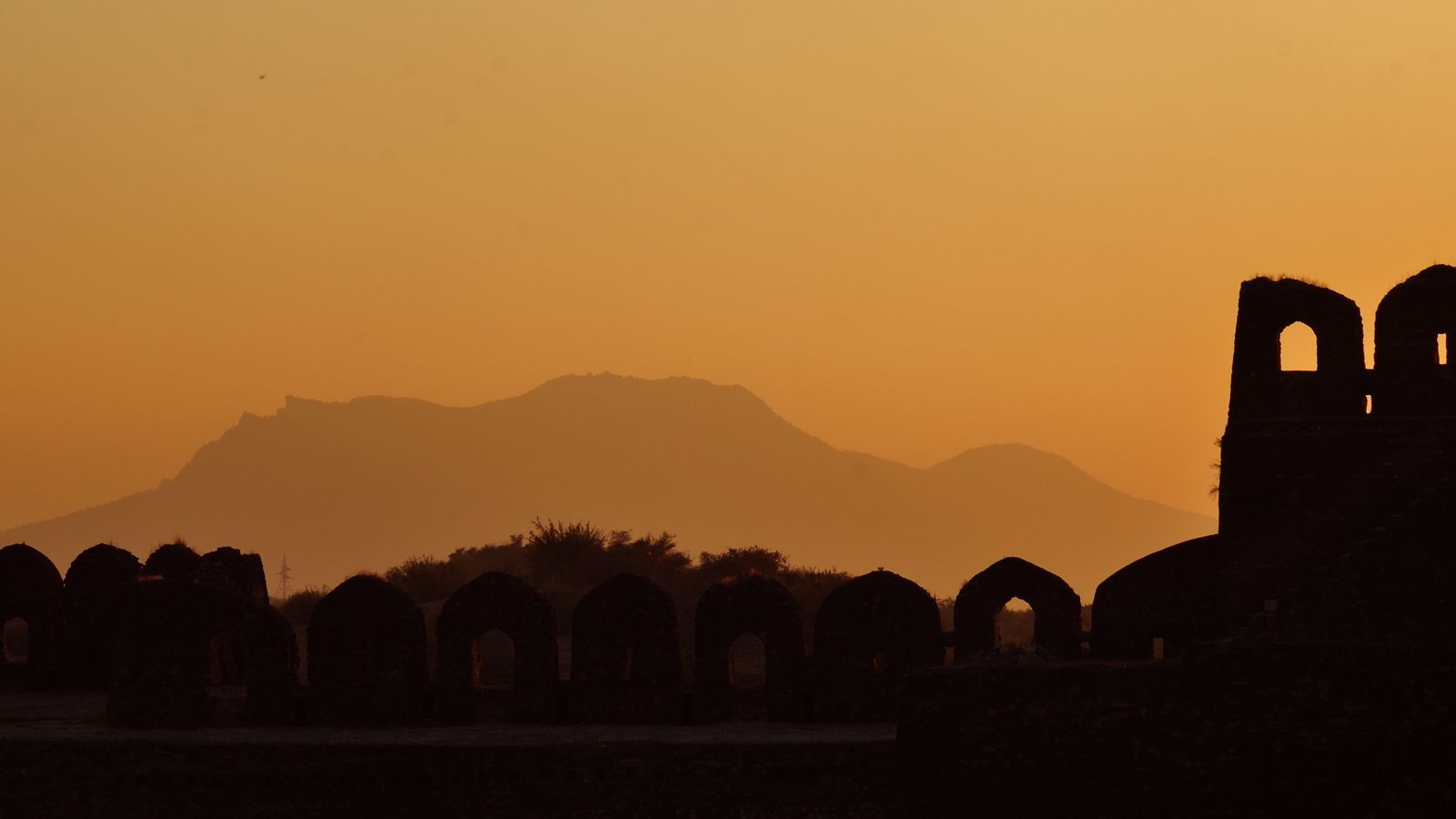 The long ridge of Tilla Jogian stands pale against an amber haze, behind the black silhouette of a row of arched battlements and a ruined tower.