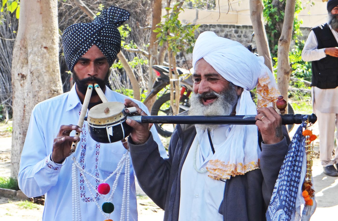 Two folk musicians in turbans under trees: a younger man plays a double flute, and an older man with a white beard, mouth open in song, holds a long-necked toomba across his chest.