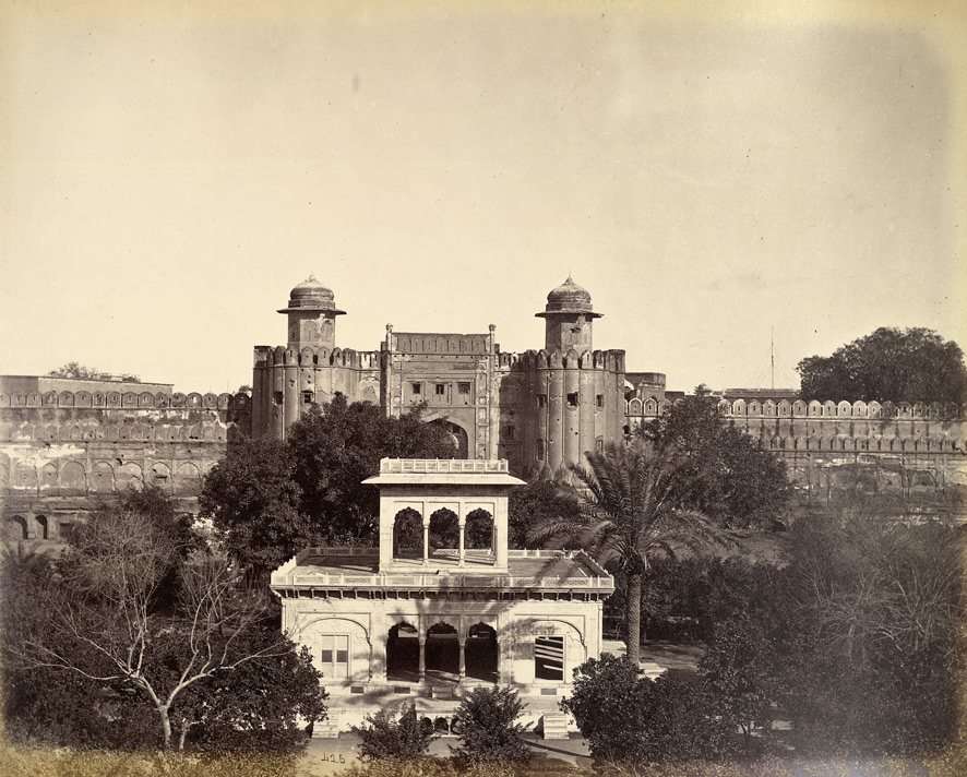 Nineteenth-century photograph of a two-storey marble pavilion among trees, with the high walls and domed towers of a fortress gateway behind it.