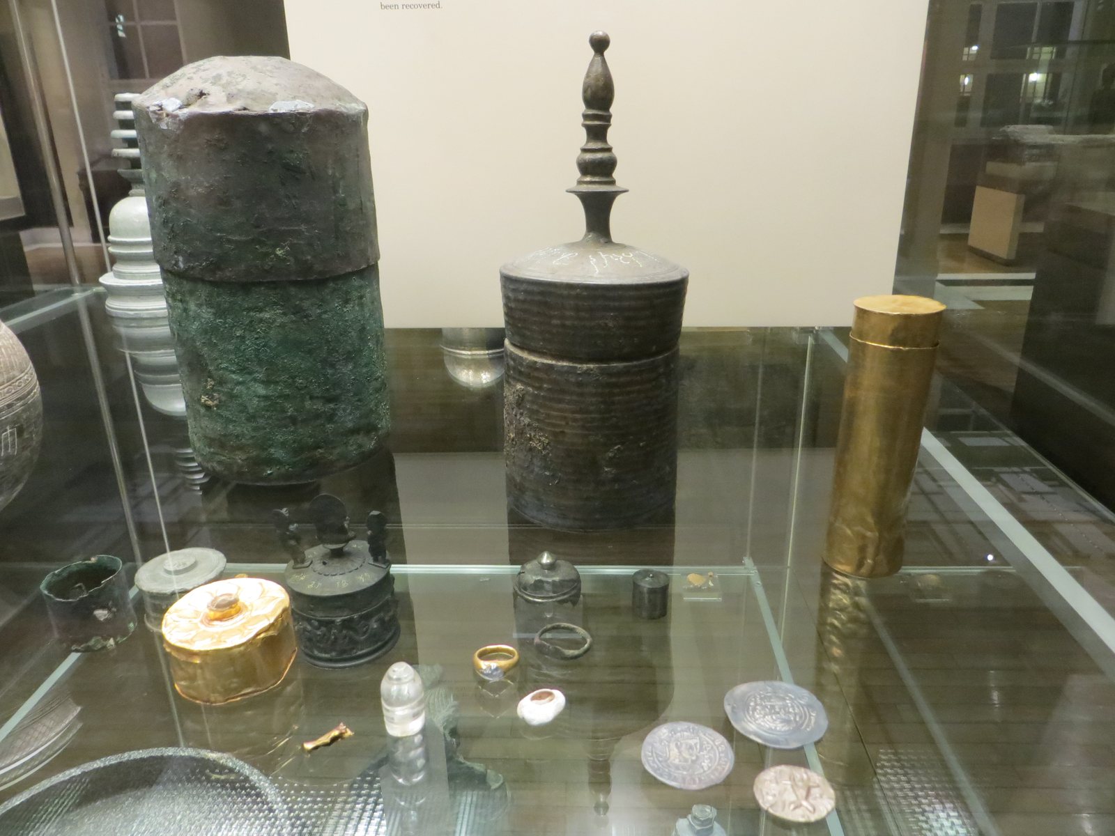 A glass museum case holding a tall corroded copper cylinder, a dark metal casket with a tall pointed finial, a gold cylinder, and in front a small gold box, a ring, a crystal and silver coins