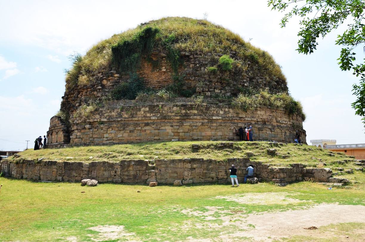 A great round stupa of dressed stone on a wide circular platform, its dome overgrown with grass and shrubs, with a few people standing at its foot for scale