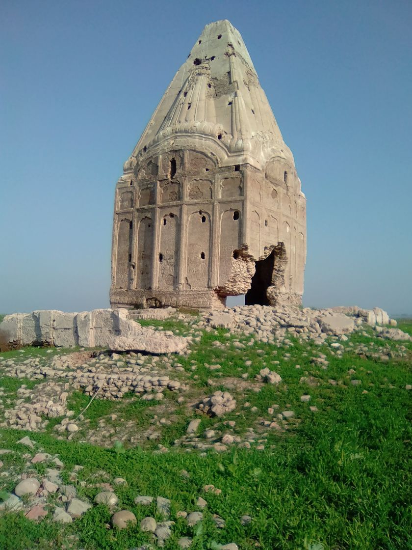 A tall, weathered temple with a pointed spire and a broken corner stands on a grassy mound strewn with stones, under a clear blue sky.