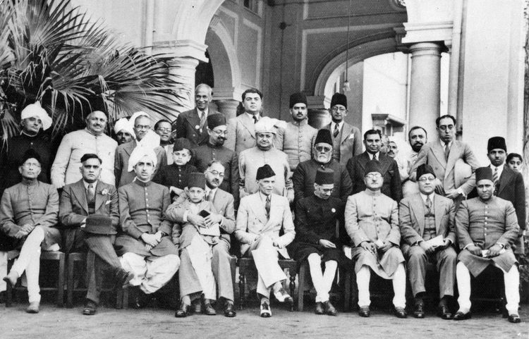 A black and white group photograph of men in sherwanis, suits and caps before an arched colonnade, one row seated and others standing behind, with Jinnah in a light suit at the centre.