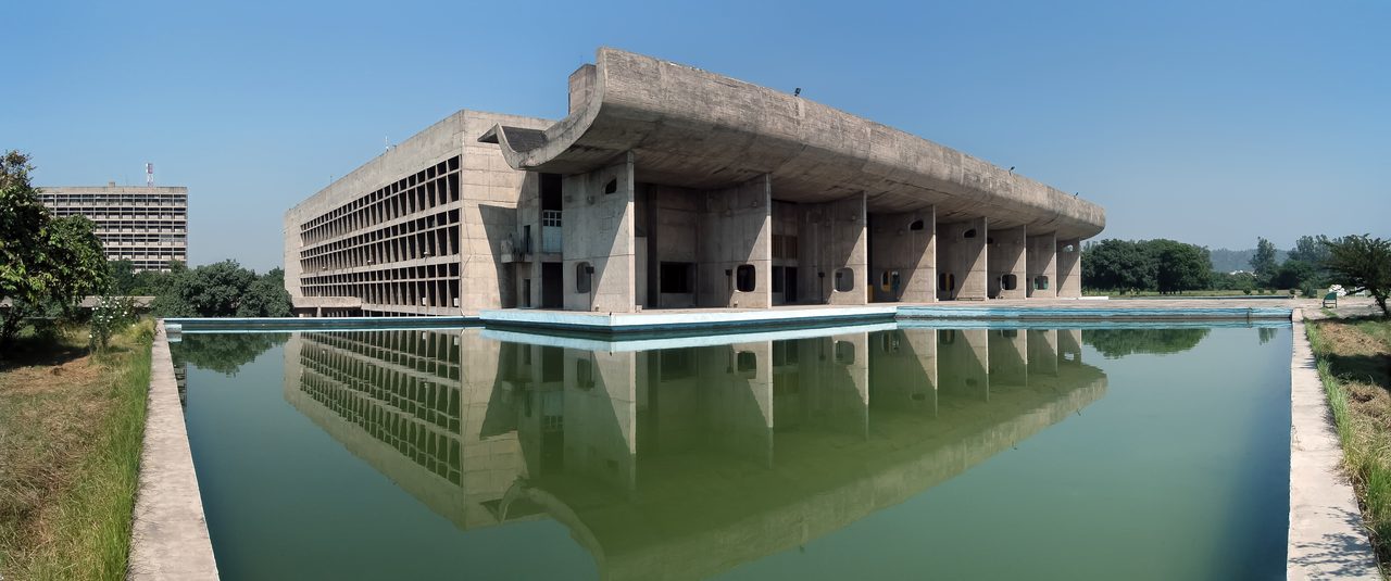 The Palace of Assembly in Chandigarh: a long concrete building with a deep upturned portico roof on tall piers, reflected in a still pool under a clear sky.