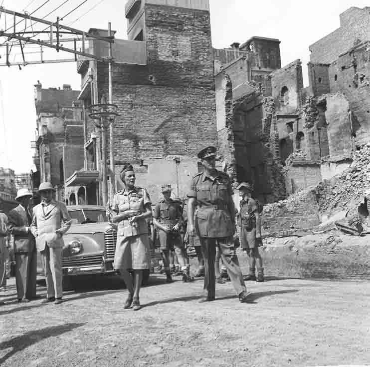 A street of gutted brick buildings with broken walls; a group of officers in uniform and a woman walk towards the camera