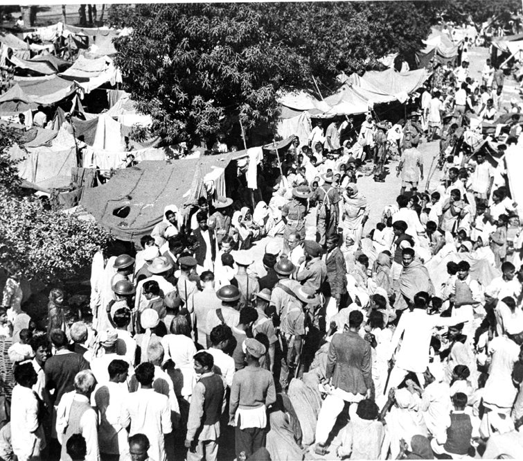 A dense crowd of people among makeshift tents and trees, with a woman in a pale uniform among them