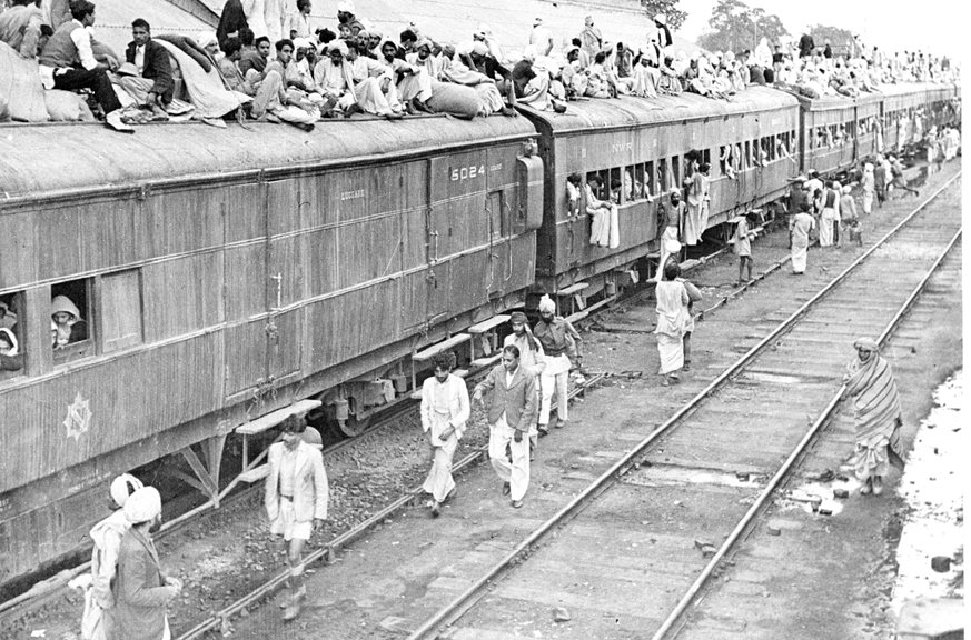 A long train of carriages with people packed inside and riding on the roofs, and more people walking beside the track