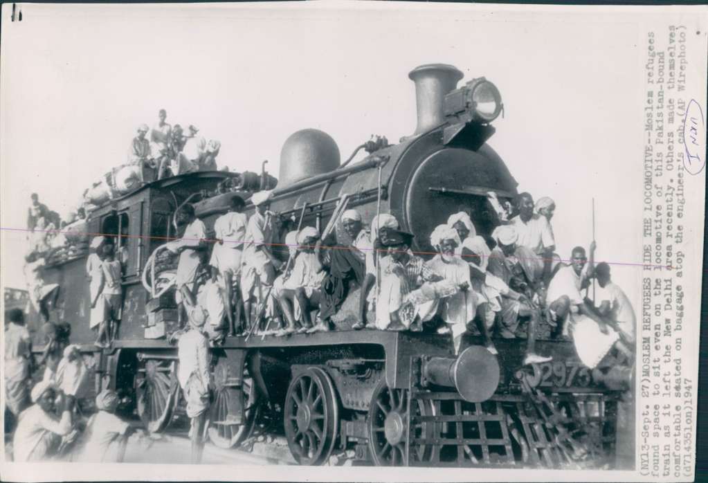 A steam locomotive crowded with men sitting on its boiler, cab roof and front, with bundles of baggage