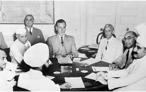 Men seated around a round table covered in papers; Mountbatten in a pale suit at the centre, Nehru in a white cap to one side, Jinnah to the other, a Sikh in a turban in the foreground with his back to the camera