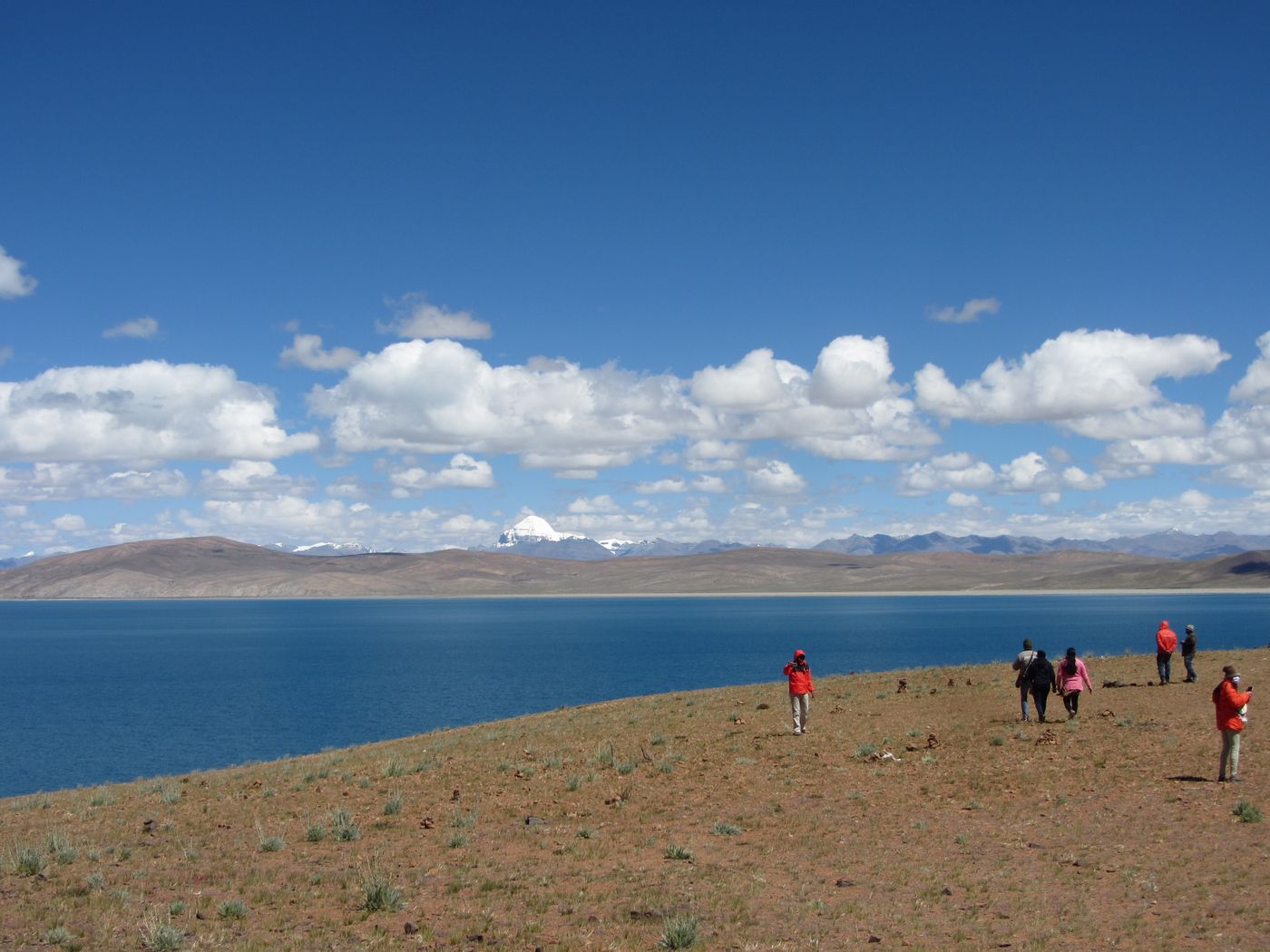 A deep blue lake under a sky of white clouds, with a low brown shore in front, a few walkers in red jackets, and a small white snow peak on the far horizon.