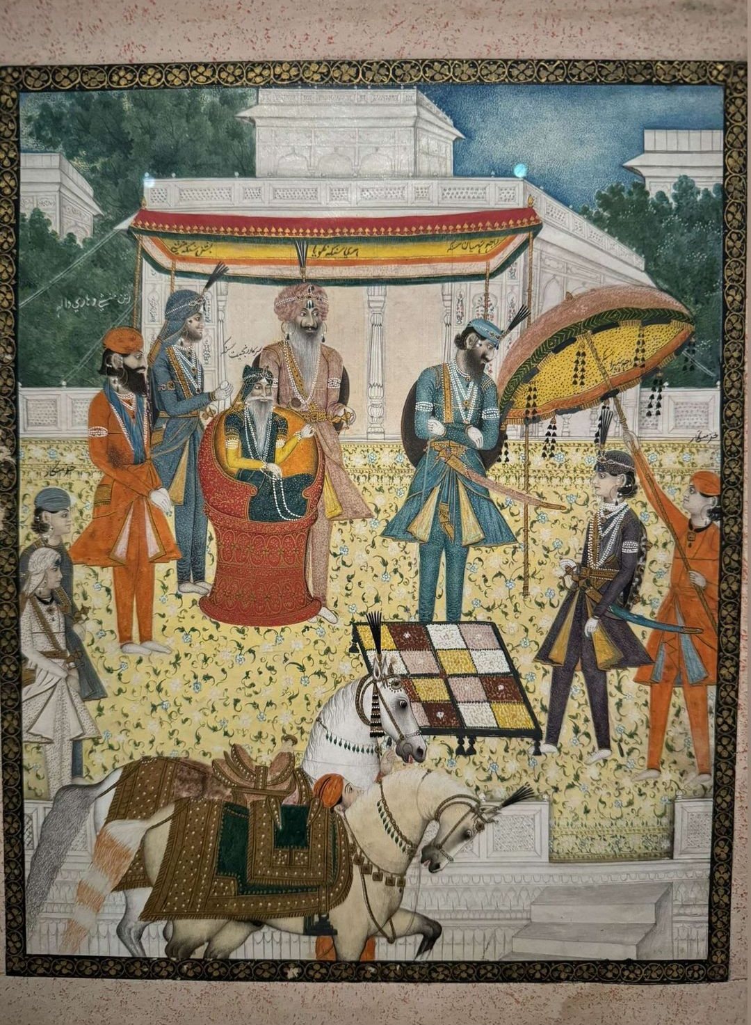 Painting of a royal terrace before a white marble pavilion: an old bearded ruler sits in a red chair under a striped canopy, courtiers in orange, blue and grey stand around him, a servant holds a yellow parasol, and two saddled white horses are led past in the foreground.