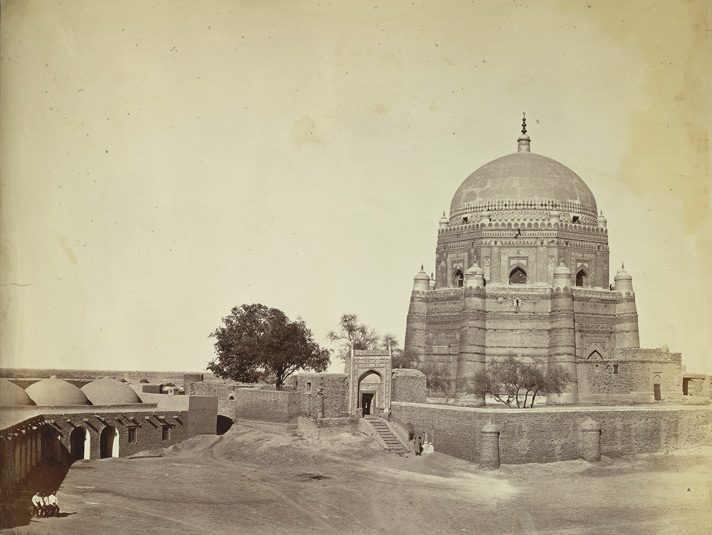 A nineteenth-century photograph of the tomb of Shah Rukn-e-Alam at Multan: a tall octagonal brick tomb with sloping corner towers and a great dome, behind a walled enclosure entered by a gateway and steps.