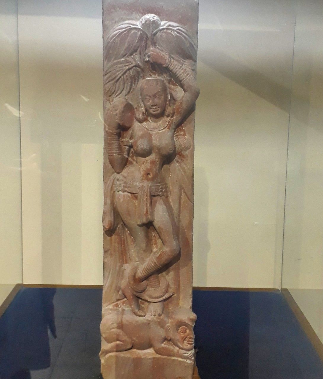 A tall red sandstone pillar carved with a woman standing cross-legged beneath a leafy tree, one arm raised to a branch, her feet on a crouching dwarf, in a glass museum case