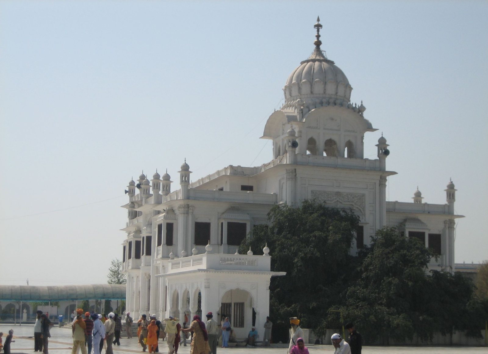A white gurdwara with a fluted dome and a long arcaded front, under a pale winter sky, with visitors walking in the courtyard
