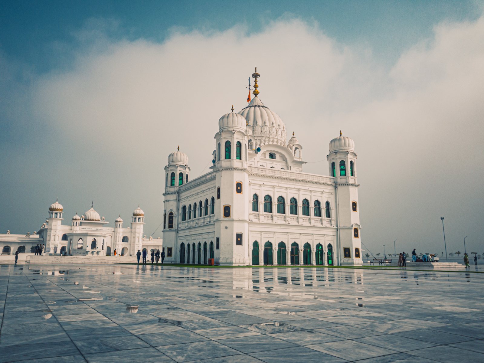 A white gurdwara with a ribbed dome, small corner domes and green-painted arches, standing at the far side of a vast, wet marble courtyard under a cloudy sky