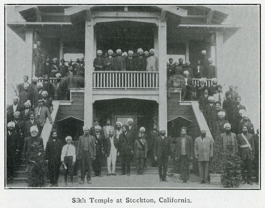 A two-storey wooden building with a deep veranda and an outside staircase, with a large crowd of turbaned men standing on the steps and in front.