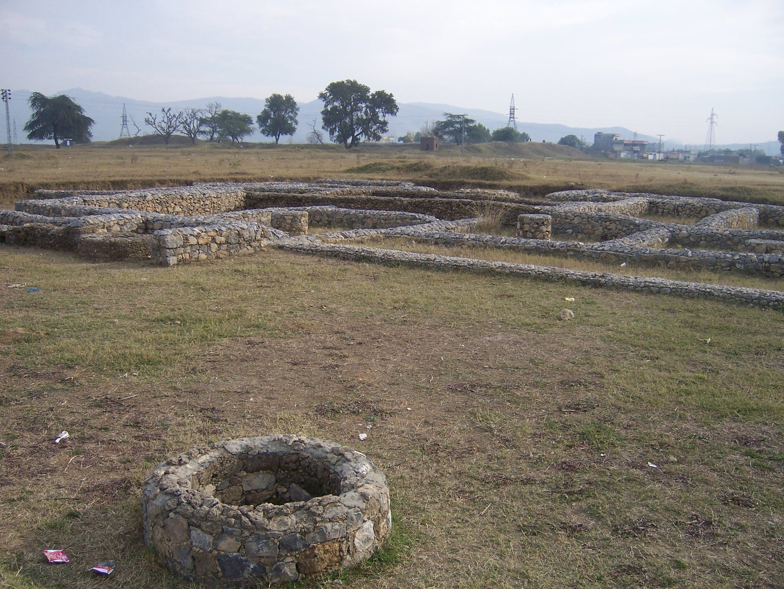 Low rubble walls of excavated houses on a flat grassy mound, with a round stone-lined well in the foreground and pylons and buildings of the modern town behind