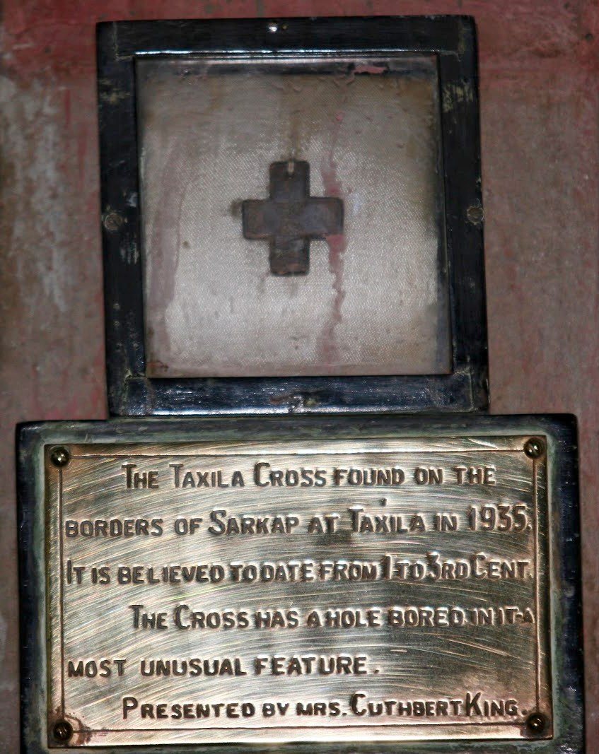 A small dark cross mounted in a glass case in a black frame, above an engraved brass plaque