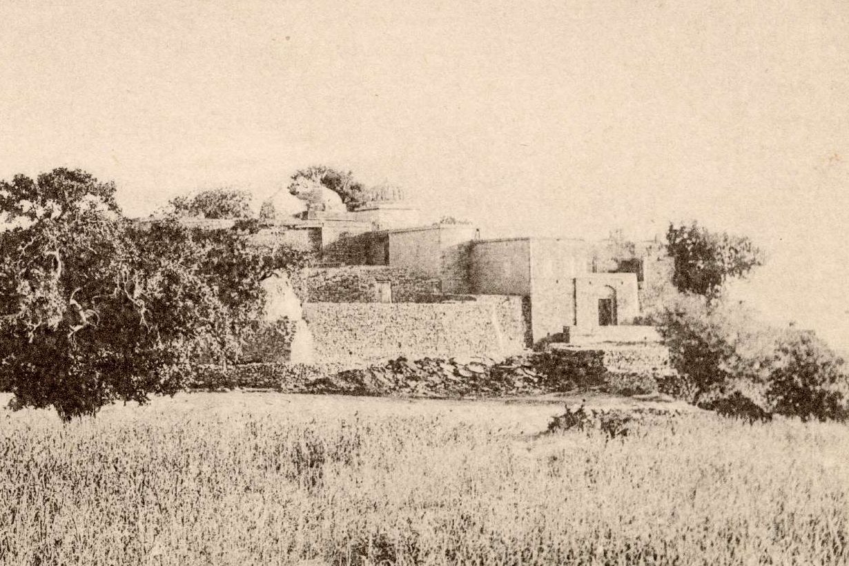 An old sepia photograph of the jogis' monastery on Tilla Jogian: flat-roofed stone buildings, walls and a small dome among trees, with a field of tall grass in front.