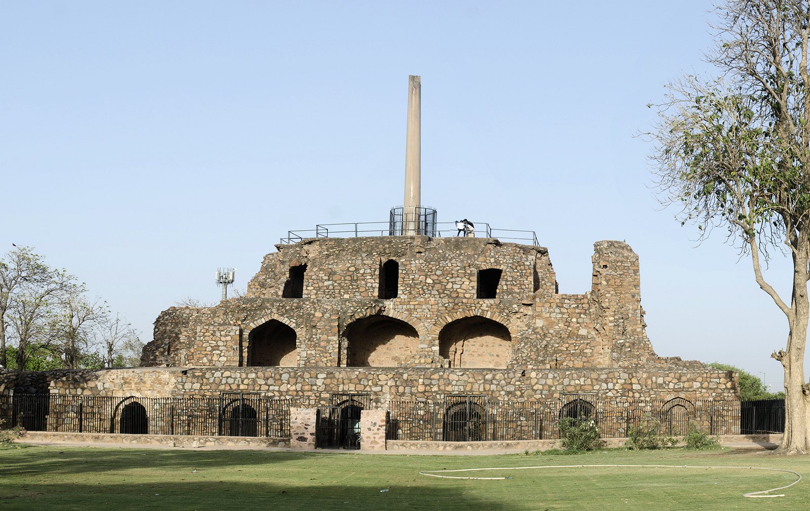 A tall, slender stone pillar rising from the top of a three-storey ruined stone building with rows of arches, on a lawn under a clear sky